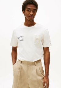 Camiseta blanca de algodón con un emblema texturizado en forma de "H" y texto bordado en azul en el lado izquierdo. Llevada con pantalones de rayas beige.