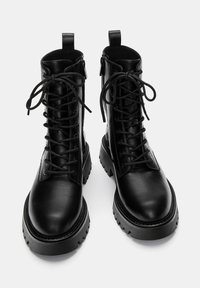 Bottes noires en cuir au niveau de la cheville avec lacets et fermetures éclair sur le côté, dotées d'une semelle en caoutchouc épaisse et de languettes pour un enfilage facile. Texture lisse.