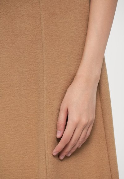 Una mano appoggiata delicatamente su un indumento di lana color cammello, con una cucitura verticale visibile su uno sfondo chiaro.