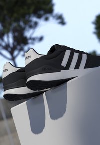 Scarpe da ginnastica Adidas nere e bianche con tre strisce bianche appoggiate su un bordo bianco all'aperto sotto un cielo sereno.