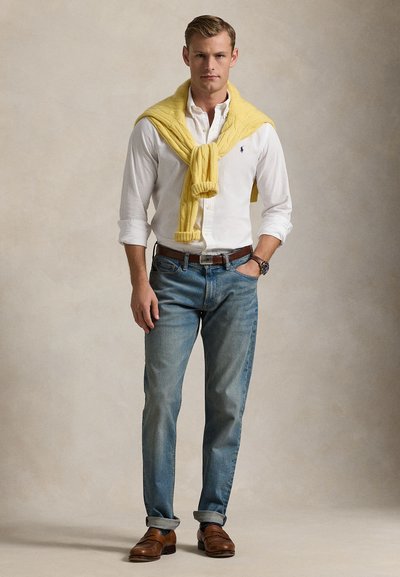 Ελαφρύ κίτρινο πλεκτό πουλόβερ επάνω από λευκό πουκάμισο με κουμπιά, σε συνδυασμό με ανοιχτό μπλε τζιν και καφέ δερμάτινα παπούτσια, παρουσιάζοντας μια κλασική casual εμφάνιση.