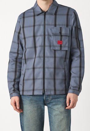 Mand iført blågråternet lynlåsjakke med frontlomme med klap og rød Hugo-mærkat, kombineret med falmede blå jeans.