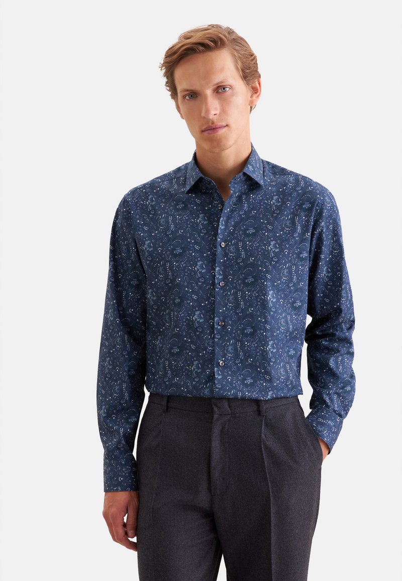 Jeune homme portant une chemise bleue à manches longues avec un motif floral subtil et un pantalon gris foncé, debout avec une main dans la poche.