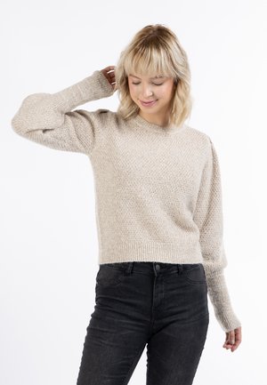 Maglione lavorato a maglia beige chiaro con un motivo testurizzato, scollo a girocollo e polsini a costine. Abbinato a jeans neri aderenti.