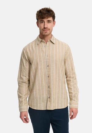 Ung mand med kort krøllet hår iført en beige langærmet skjorte med hvide lodrette striber og mørke bukser, stående med fronten vendt mod kameraet.