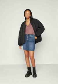 Μαύρο bomber jacket, μωβ ριμπ τοπ, τζιν μίνι φούστα με κουμπιά, μαύρες μπότες με κορδόνια, σε στάση όρθια σε ουδέτερο φόντο.