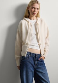 Cardigan con cappuccio di un beige chiaro realizzato in materiale morbido, caratterizzato da una chiusura frontale con zip e polsini a costine, indossato sopra un top bianco e jeans blu.