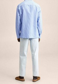 Camicia azzurra a righe con bottoni e maniche lunghe; abbinata a pantaloni di jeans chiari. Le scarpe slip-on marroni completano l'outfit.