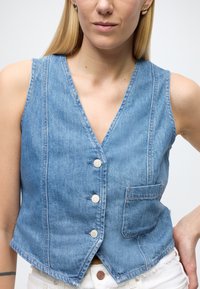 Jeans brezrokavnik svetlo modre barve, brez rokavov, z V-izrezom, petimi gumbi, en žep spredaj, neobdelan rob, mehka tekstura in šivani detajli.