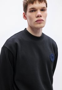 Μαύρο φούτερ με στρογγυλό κεντημένο μπλε περιλαίμιο στο στήθος, με υφασματικό σχέδιο και ριμπ λαιμόκοψη και μάνικες.