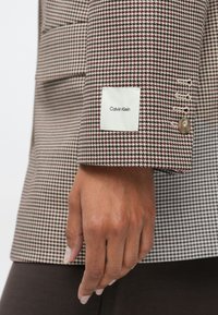 Main avec des ongles naturels à côté de la manche d'un blazer pied-de-poule marron et blanc avec une étiquette Calvin Klein et des boutons beiges.
