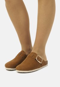 Chaussures slip-on en cuir suédé marron à bout rond, dotées d'une grande boucle métallique et d'un accent en liège texturé sur le côté.