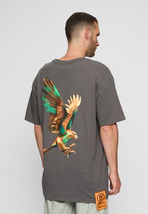 Grå oversized t-skjorte med et fargerikt ørnegraphic på baksiden, med detaljerte vinger og klør, samt et oransje merke på siden.