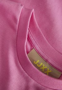 Tissu en coton rose avec un col côtelé. L'étiquette présente le texte "JJXX" en jaune. Texture douce, finition lisse et design décontracté.