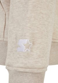 Lys beige sweatshirt-stoff med en myk tekstur. Har en brodert hvit logo med en geometrisk form og stjerne på ermet.