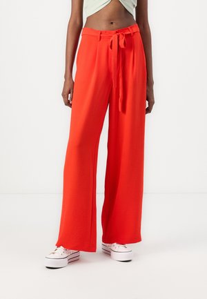 Mujer con pantalones anchos de color naranja brillante con cinturón atado, combinados con zapatillas blancas de plataforma sobre un fondo liso.