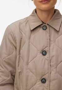 Inimese lähivõte, kes kannab beeži värvi tikitud jopet, millel on suured mustad nööbid ja terav krae.