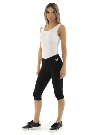 Une femme porte un haut blanc sans manches, un legging noir qui arrive aux genoux et des baskets noires sur un fond blanc uni.