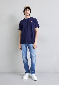 Ναυτικό βαμβακερό T-shirt με κόκκινο λογότυπο, συνδυασμένο με ανοιχτόχρωμο μπλε τζιν και λευκά αθλητικά παπούτσια. Μοντέλο που φοριέται καπέλο σε τόνο μπεζ, στέκεται μπροστά από έναν ουδέτερο τοίχο.