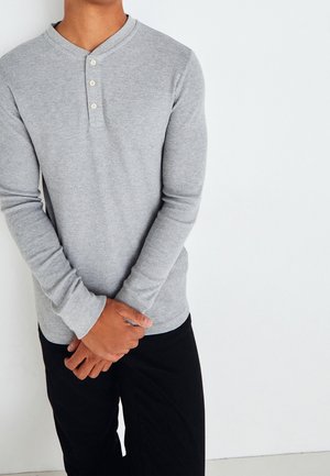 Uomo che indossa una maglia henley grigia aderente a maniche lunghe con tre bottoni, mani intrecciate davanti, pantaloni neri, su sfondo bianco.