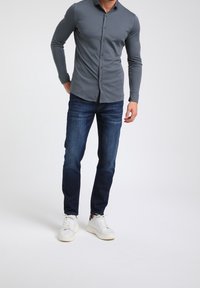 Camisa gris de manga larga con botones combinada con vaqueros azul oscuro y zapatillas blancas, mostrando un diseño ajustado y una textura suave.