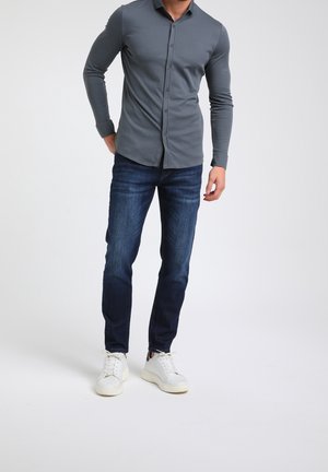 Graues Langarm-Hemd mit Knöpfen, kombiniert mit dunkelblauen Jeans und weißen Sneakers, zeigt ein tailliertes Design und eine glatte Textur.