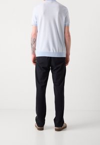 Polo de punto azul claro de mangas cortas con un cuello y puños acanalados. Combinado con pantalones negros y zapatos marrones. La vista trasera muestra un tatuaje en el brazo.