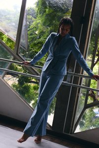 Persoană în costum albastru, desculță, echilibrându-se pe un picior în timp ce ține o balustradă într-o încăpere cu ferestre mari înclinate și copaci verzi vizibili afară.