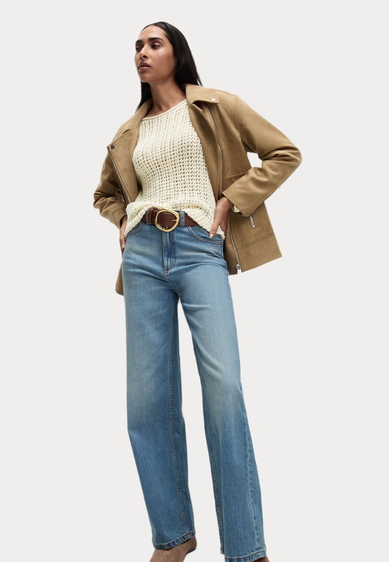 Tan leren jack over een crèmekleurige gebreide trui, gecombineerd met lichtblauwe wijde jeans. Bruine riem met grote ronde gesp.