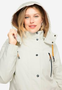 Vaaleanbeige takki, jossa on huppu, suuret napit ja vetoketjulliset taskut. Takissa on elastista yksityiskohtaa hihansuissa ja värikäs naru korostuksena.