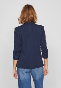 Blazer marine avec une silhouette structurée, des manches longues et des épaulettes. Tissu doux avec une texture lisse. Associé à un jean délavé.
