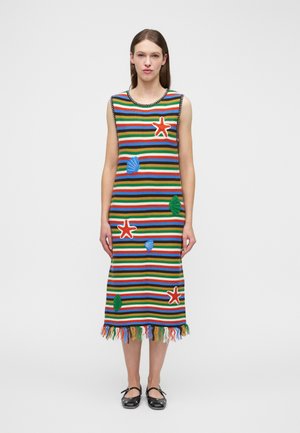 Femme portant une robe rayée sans manches, longueur au genou, ornée de patchs à thème marin et d'un ourlet à franges, debout devant un fond blanc uni.