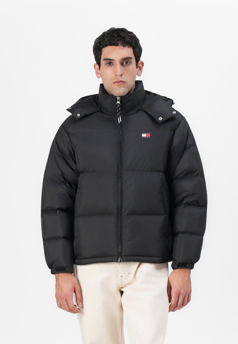 Schwarze Pufferjacke mit hohem Kragen, Reißverschluss und Kordelzugkapuze. Verfügt über eine Steppnaht und ein kleines Logo auf der Brust.