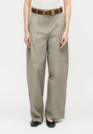 Ženska, ki nosi bež hlače z visokim pasom in širokimi nogavicami s sprednjimi šivi, rjavi usnjeni pas, belo majico in črne čevlje s kvadratnim prstom.