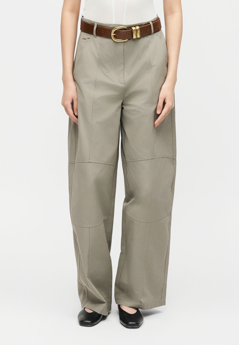 Γυναίκα που φοράει μπεζ παντελόνι με ψηλή μέση και φαρδιά μπατζάκια με λεπτομέρειες ραφής μπροστά, καφέ δερμάτινη ζώνη, λευκό τοπ και μαύρα παπούτσια με τετράγωνη μύτη.