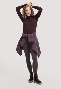 Burgunderrotes Langarmoberteil, kombiniert mit dunkelgrauen Leggings und einer lila Fleecejacke, die um die Taille gebunden ist. Braune Stiefeletten mit einer strukturierten Sohle.