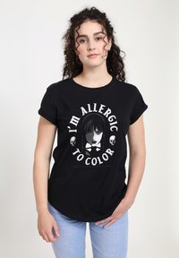 Ung kvinde med krøllet hår bærer en sort t-shirt med teksten "Jeg er allergisk overfor farve" og en illustration af et monocromt pigeface og kranier.