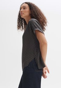 Camicia a maniche corte blu navy con un motivo geometrico in crema e oro, caratterizzata da una vestibilità rilassata e spacchi laterali, abbinata a jeans di denim scuro.