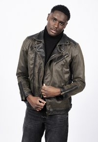 Młody mężczyzna poprawiający ciemnobrązową skórzaną kurtkę z detalami na suwak, ubrany w czarny golf i czarne dżinsy na białym tle.