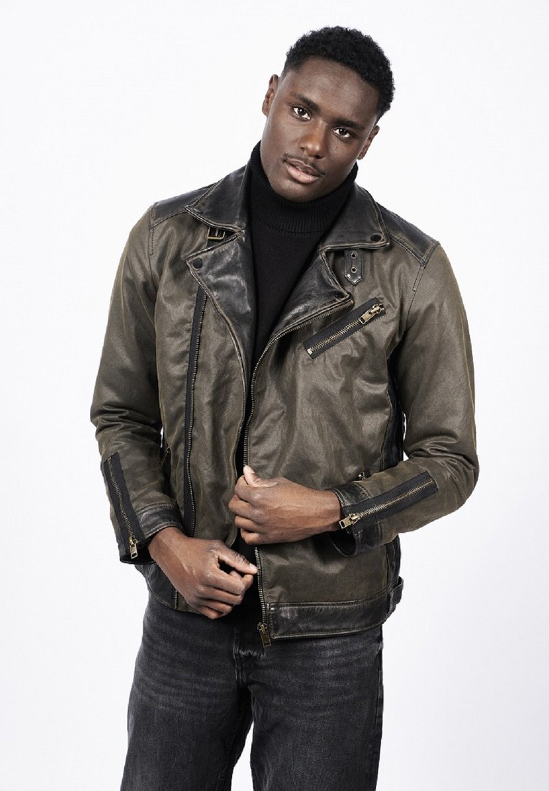 Młody mężczyzna poprawiający ciemnobrązową skórzaną kurtkę z detalami na suwak, ubrany w czarny golf i czarne dżinsy na białym tle.