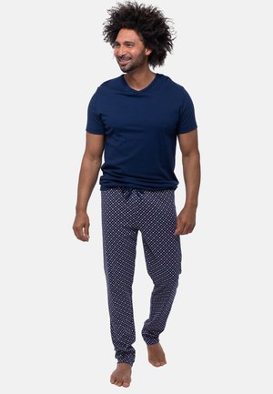 Man met krullend haar die een marineblauw T-shirt en patroonpyjamabroek draagt, blootsvoets loopt tegen een effen achtergrond.