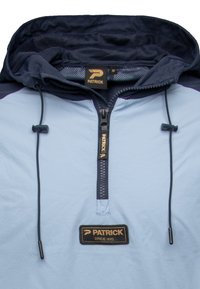 Chaqueta de pullover azul claro con capucha azul marino. Cuenta con un cierre frontal con cremallera y una etiqueta de tela con la marca "PATRICK". Incluye un cordón ajustable.