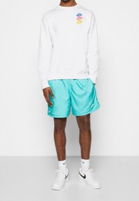 Bílá mikina s kapucí s vícebarevnými logy Nike na hrudi, spárovaná s tyrkysovými šortkami, bílými ponožkami a černými teniskami Nike.