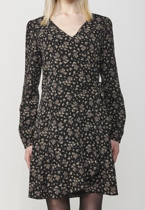 Zwarte lange mouw wikkeljurk met een klein beige bloemmotief, V-hals en licht uitlopende rok, gedragen over zwarte panty's.