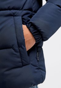 Marineblå dunjakke med quiltet design, med elastisk mansjett og sidelomme. Logotagg synlig nær lommen.