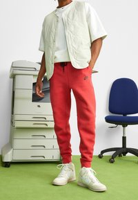 Nike fodrad väst i ljusgrön, vit t-shirt, röda svettbyxor med logotyp och krämfärgade sneakers. Står bredvid en kopieringsmaskin.