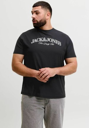 Mann mit Bart, der ein schwarzes "JACK&JONES For Daily Use" T-Shirt und graue Hose trägt, steht mit verschränkten Händen und schaut zur Seite.