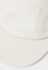 Beige cotton cap with a curved brim, featuring textured fabric and visible stitching lines. Simple design with no prominent patterns or accents.
