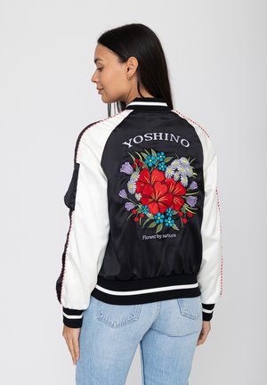 Frau mit langen dunklen Haaren, die eine schwarz-weiße Jacke mit bunter Blumenstickerei und dem Schriftzug "YOSHINO Flower by nature" auf dem Rücken trägt.
