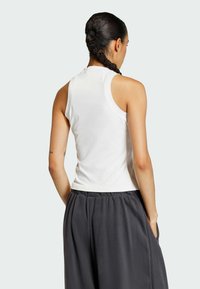 Biela bezrukávová blúzka s okrúhlym výstrihom, skombinovaná s voľnými tmavosivými nohavicami so bočnými vreckami a plistenou textúrou. Zobrazený zadný pohľad.
